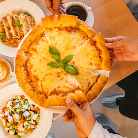 Person holding a artisan fully baked pizza with a side of salad and coffee on a wooden table by Bake N More bakery manufacturer in Dubai